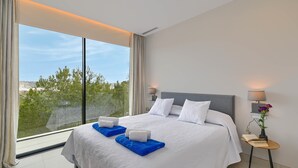 Une chambre d’hôtel moderne avec un grand lit, une table de chevet, une lampe et une vue sur la verdure grâce à de grandes fenêtres.