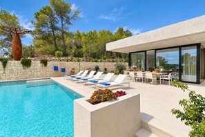 A modern outdoor pool area with lounge chairs, a dining table, and a view of trees and hills.
