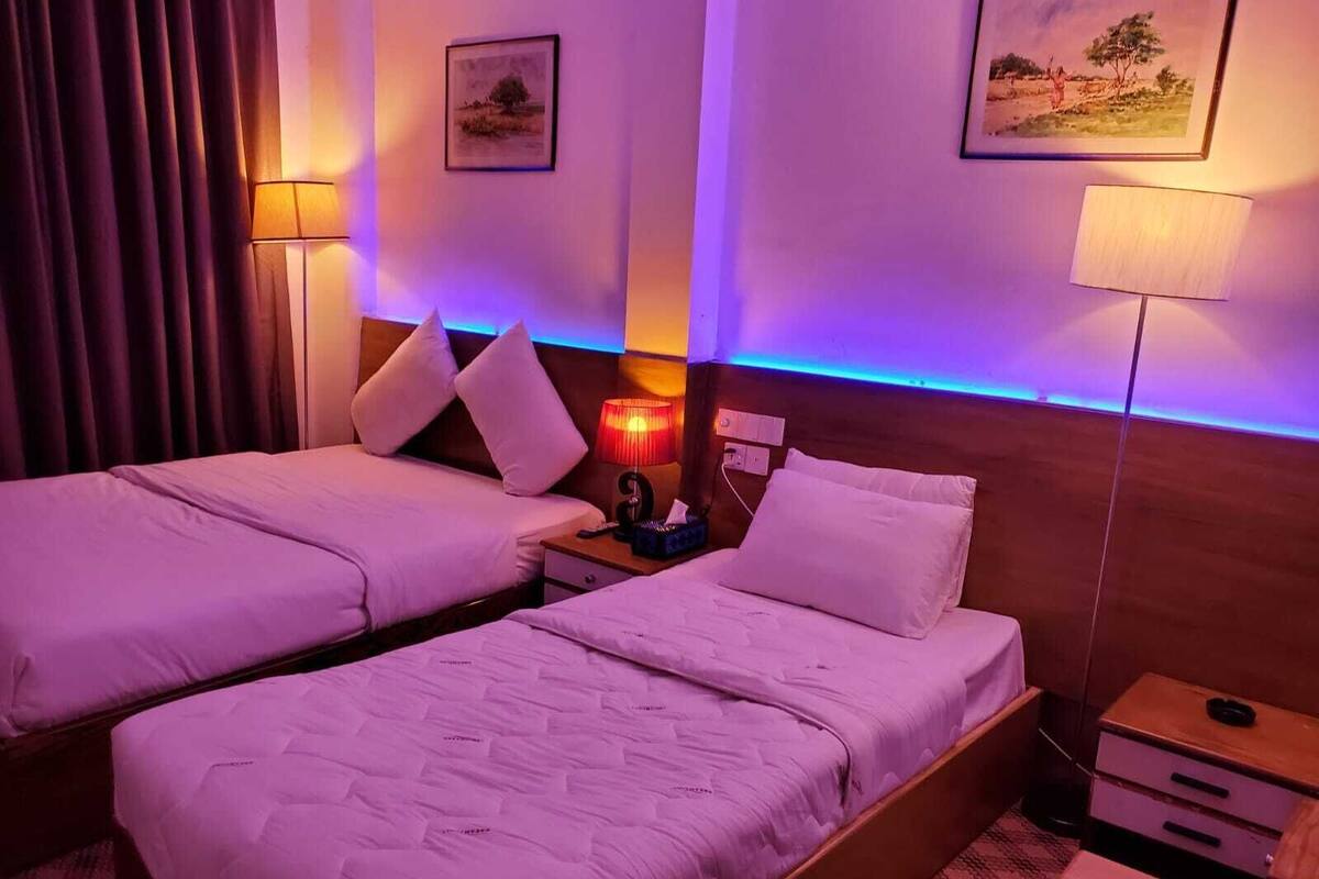 A hotel room with two beds, each with white bedding and pillows. There are two wall-mounted lamps, one with a red light and another with a white light. The room has a wooden headboard and a small wooden nightstand with a lamp and a phone.