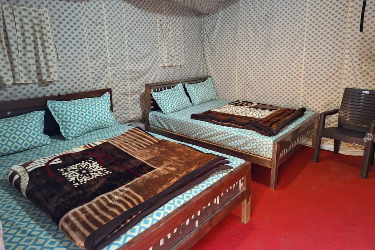 A room with two beds, each with patterned bedding and a wooden headboard. There is a red carpet on the floor and a chair in the corner.