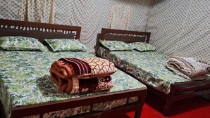 A room with two beds, each with a wooden frame and floral-patterned bedding. There are rolled blankets on the beds, and a red cylindrical object is visible on the floor.