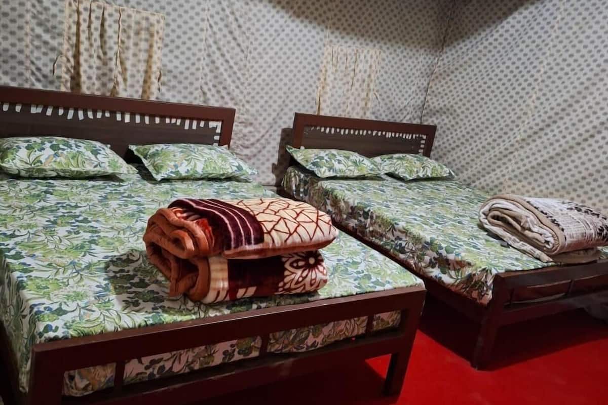 A room with two beds, each with a wooden frame and floral-patterned bedding. There are rolled blankets on the beds, and a red cylindrical object is visible on the floor.