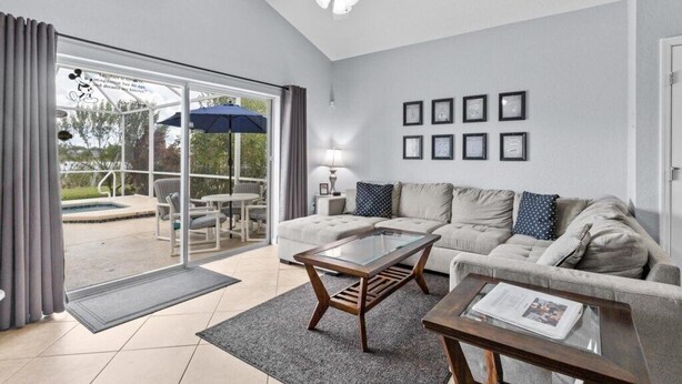 Ein Wohnzimmer mit Sofa, Couchtisch und Deckenventilator. Ein Schiebetürfenster führt zu einem Außenbereich mit Pool und Terrassenmöbeln.