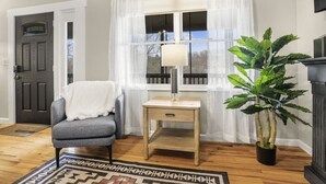 Une pièce avec un plancher en bois, un fauteuil gris, une petite table d’appoint en bois sur laquelle se trouve une lampe, une plante en pot et une vue par une fenêtre ornée de rideaux.