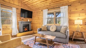 L’intérieur chaleureux d’un chalet avec une cheminée, un canapé, un téléviseur et un plafond en bois.