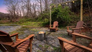 Une terrasse en bois, dotée de chaises Adirondack et d’un foyer, entourée d’arbres et agrémentée d’une pergola en bois illuminée de guirlandes lumineuses.