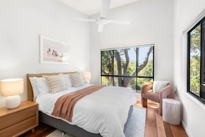 Quarto com cama, mesas de cabeceira, ventilador de teto, janela com vista para árvores e uma cadeira.