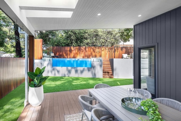 A modern outdoor patio with a wooden deck, a pool, and a dining area with chairs and a table.