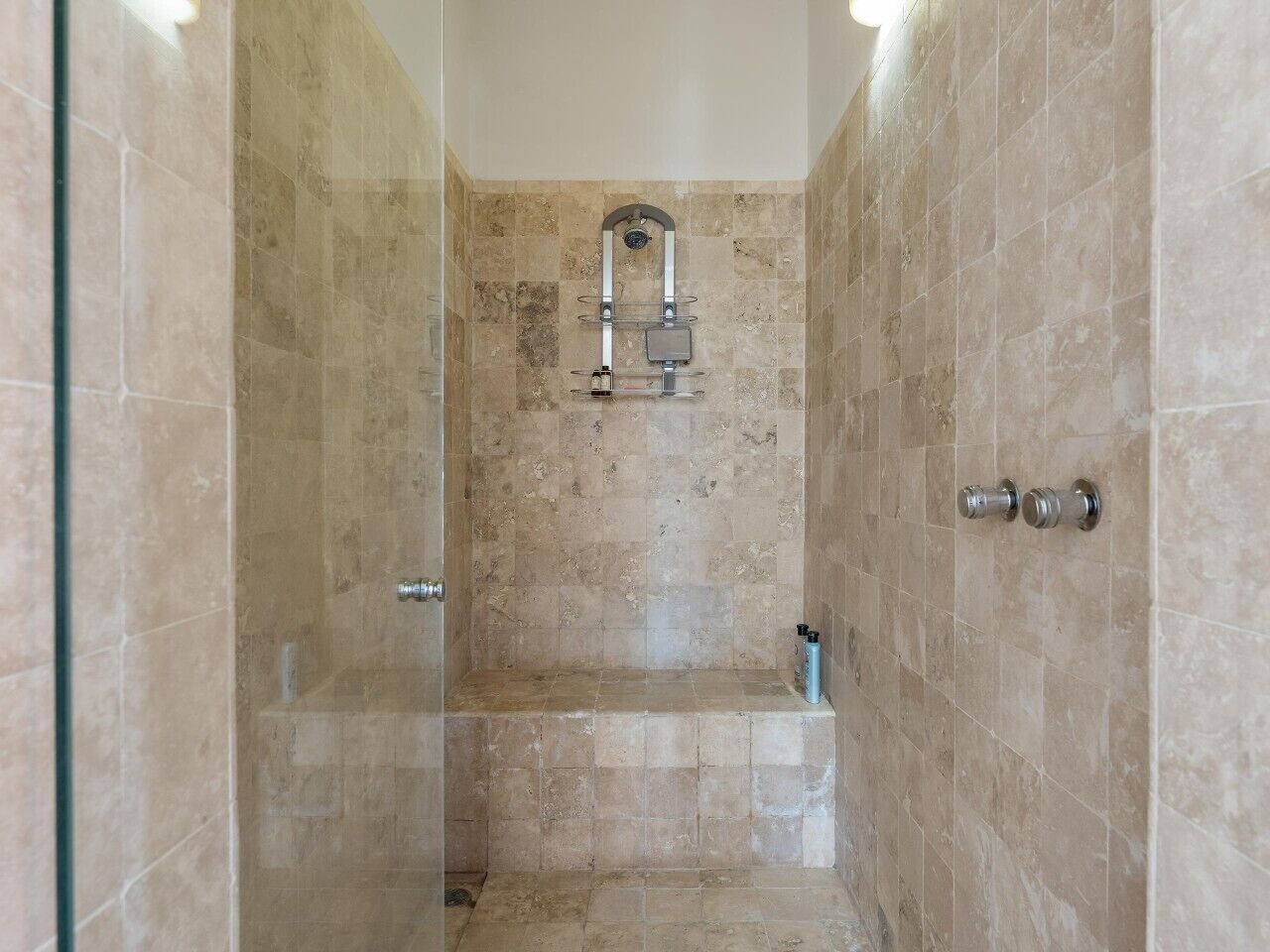 Zona de ducha con cerramiento de vidrio, azulejos color beige, un cabezal de ducha y un estante con artículos de aseo.
