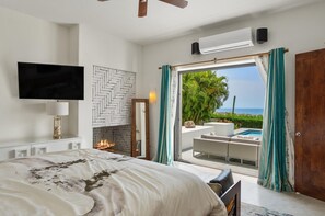A bedroom with a bed, a TV, a fireplace, and a view of the ocean.