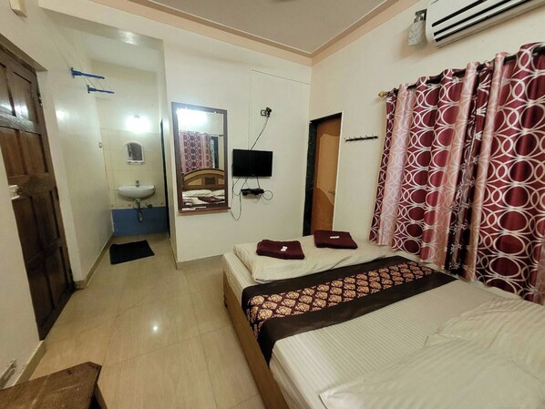 Habitación de hotel con cama, mesita de noche, espejo, televisor, ventana con cortinas y baño con lavamanos y espejo.