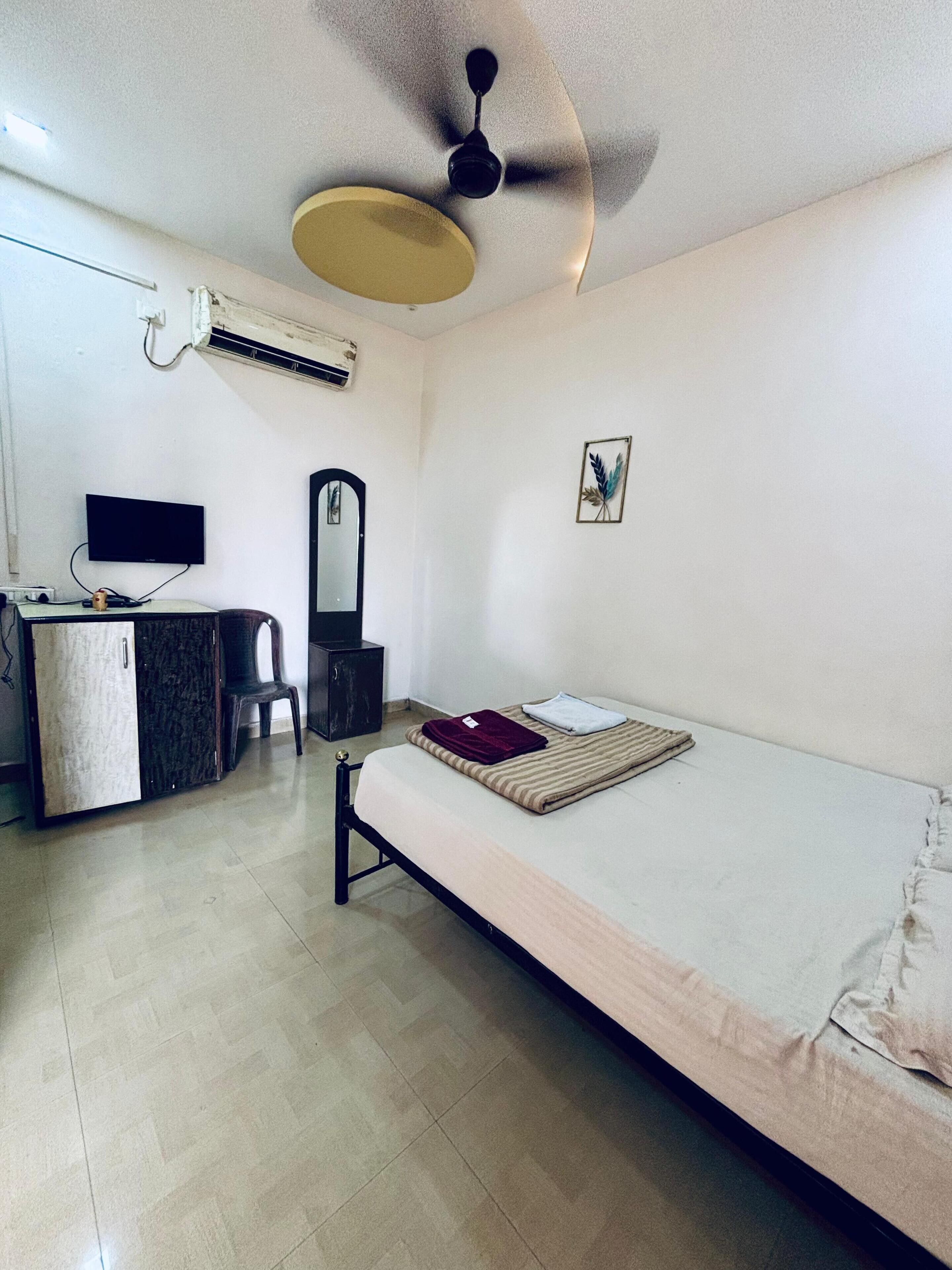 Une chambre à coucher comprenant un lit, un bureau, une chaise, un miroir et un ventilateur de plafond.