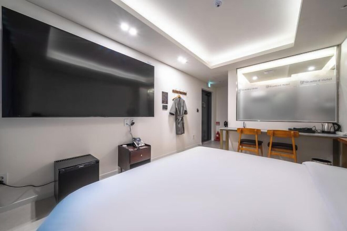 A modern hotel room with a large bed, a flat-screen TV, a desk with a chair, and a whiteboard.