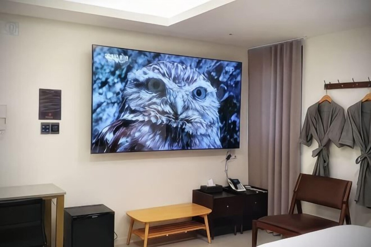 A hotel room with a large wall-mounted TV, a bed, a desk, a chair, and a wardrobe.
