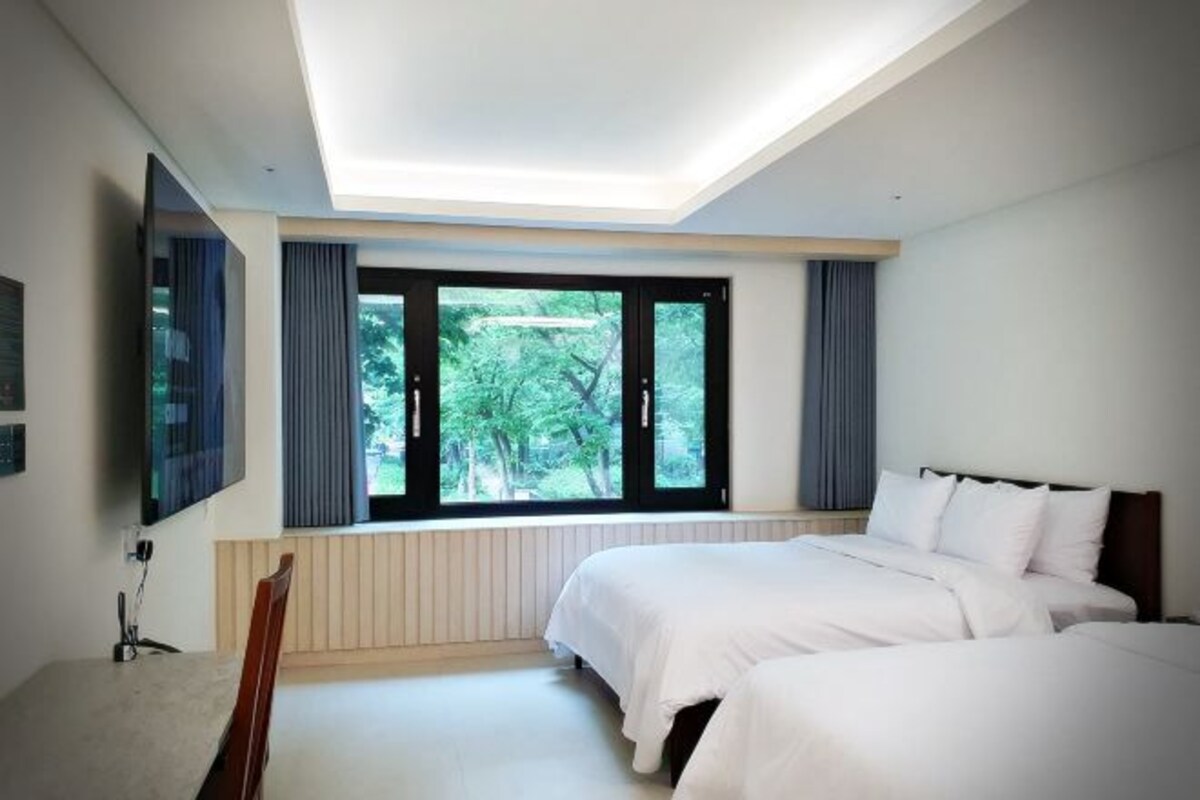 A bedroom with a large window, a bed, a chair, and a flat-screen TV mounted on the wall.