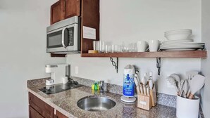 A kitchen with a granite countertop, stainless steel sink, wooden cabinets, a microwave, and a coffee maker. - Pelican Studio w/ Private Patio • Pompano (Pompano Beach)