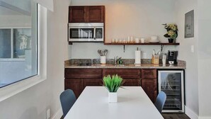 Una cocina moderna con armarios de madera oscura, un microondas integrado, una cafetera y un refrigerador pequeño. Hay un comedor blanco con dos sillas y una planta en maceta encima.