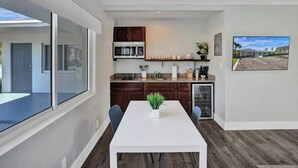 Una cocina moderna con armarios de madera oscura, una encimera blanca y un microondas integrado. Hay una zona de comedor con una mesa y sillas blancas, y una planta en maceta sobre la mesa.
