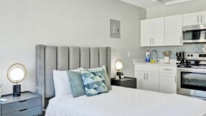 Una habitación de hotel moderna con una cama, mesitas de noche, un espejo, una kitchenette con microondas y horno, y un interruptor de luz en la pared.
