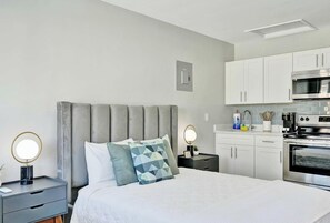 Moderni hotellihuone, jossa on sänky, yöpöydät, yölamppu, peili sekä keittiöalue, jossa on kaappeja ja kodinkoneita.