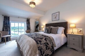 Ein Schlafzimmer mit einem großen Bett, einem Nachttisch, einem Sessel, einem Fenster mit Vorhängen und einer Deckenleuchte.