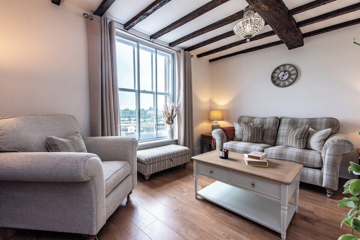 A living room with a sofa, armchair, and a coffee table. There is a window with curtains, a lamp, and a clock on the wall.