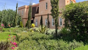 A large, multi-story building with a flat roof and a symmetrical facade, surrounded by lush greenery and flowering plants. -  Relax in the beauty of a saffron farm in the foothills of Atlas Mountains  (Marrakech)