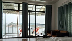 A hotel room with a large sliding glass door leading to a balcony with a view of a lake. There is a bed, a small table, and two orange chairs on the balcony. - Rangadip Resort Rangamati (Rangamati Sadar)