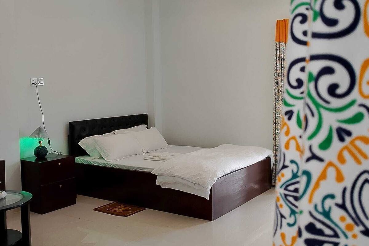 A bedroom with a bed, nightstands, a lamp, a ceiling fan, and a patterned curtain.