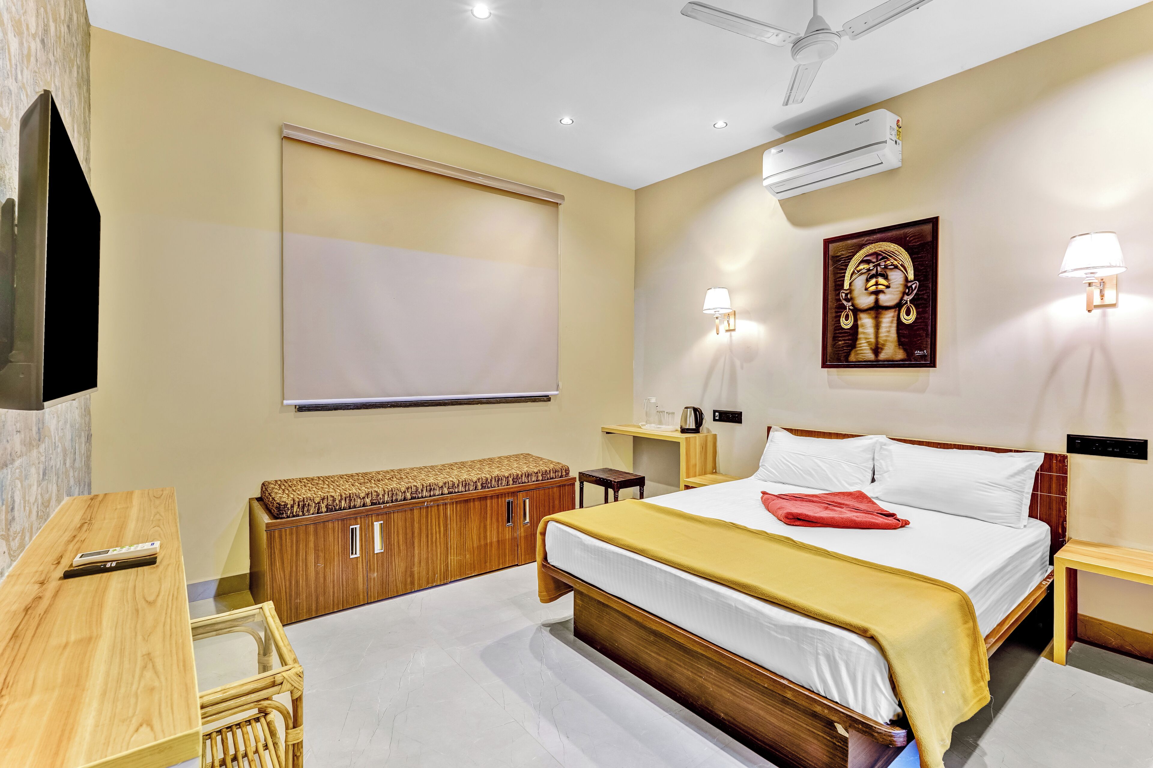 A hotel room with a bed, bedside tables, a wooden bench, a TV, and an air conditioning unit.