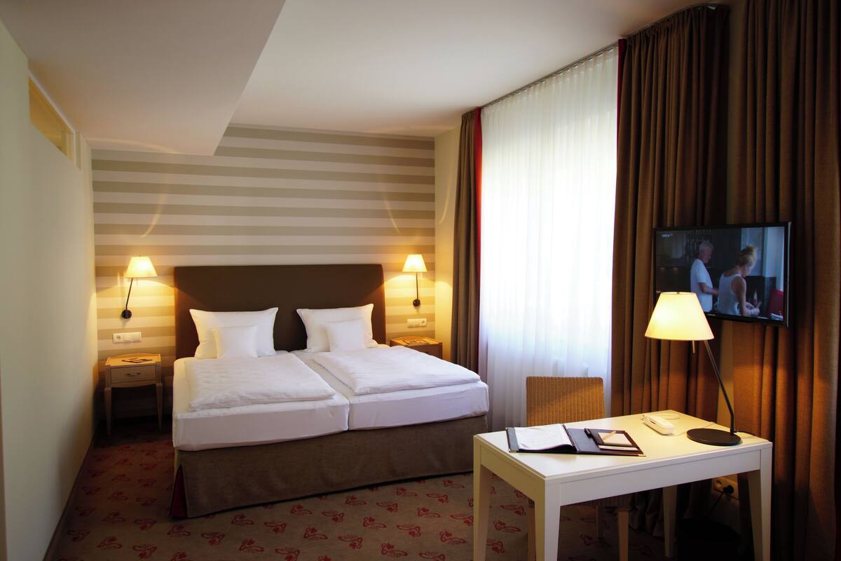 A hotel room with a double bed, two bedside lamps, a TV on the wall, a desk with a lamp, and a patterned carpet.