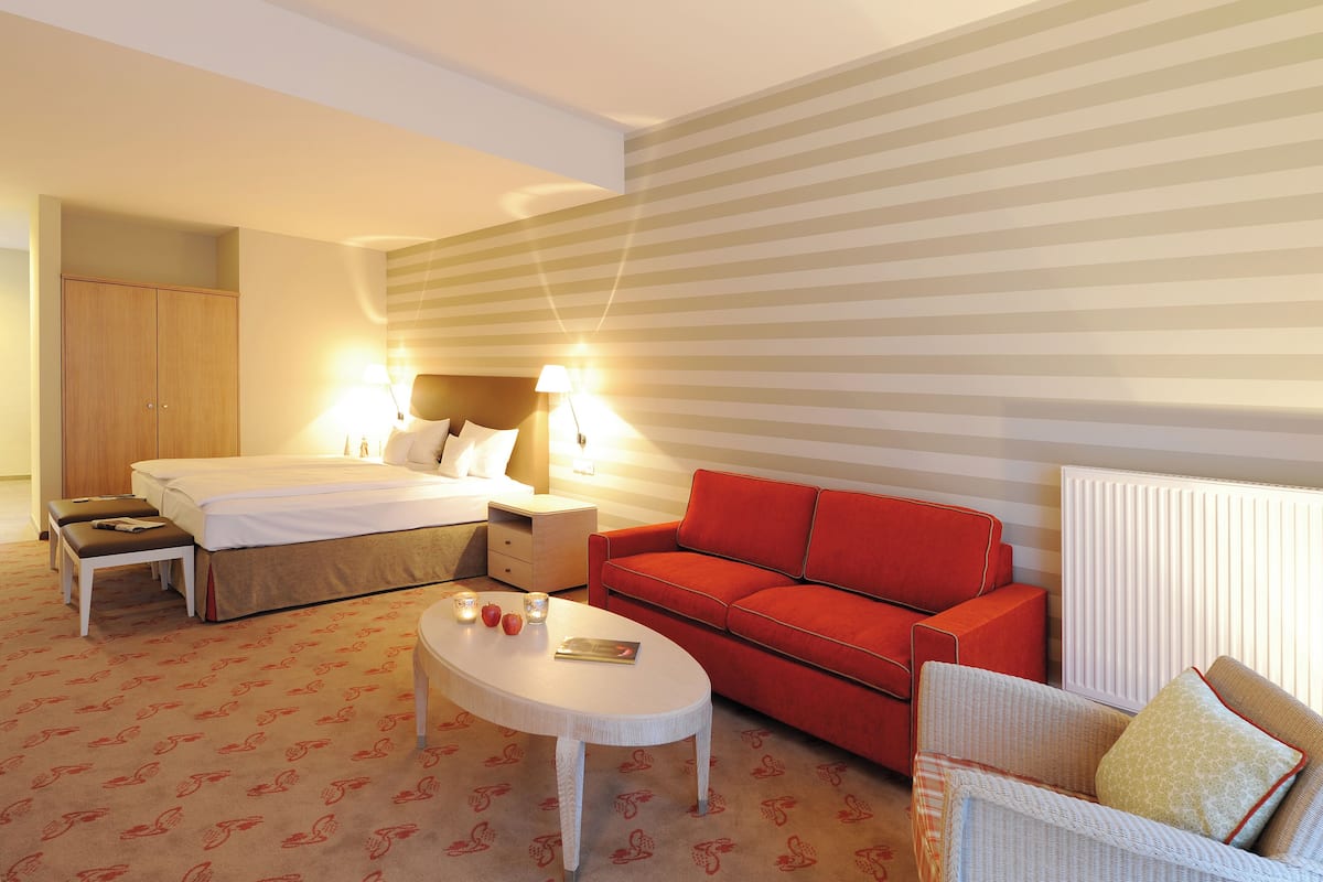 A hotel room with a bed, a red sofa, a white armchair, a small table, and a nightstand with a lamp.