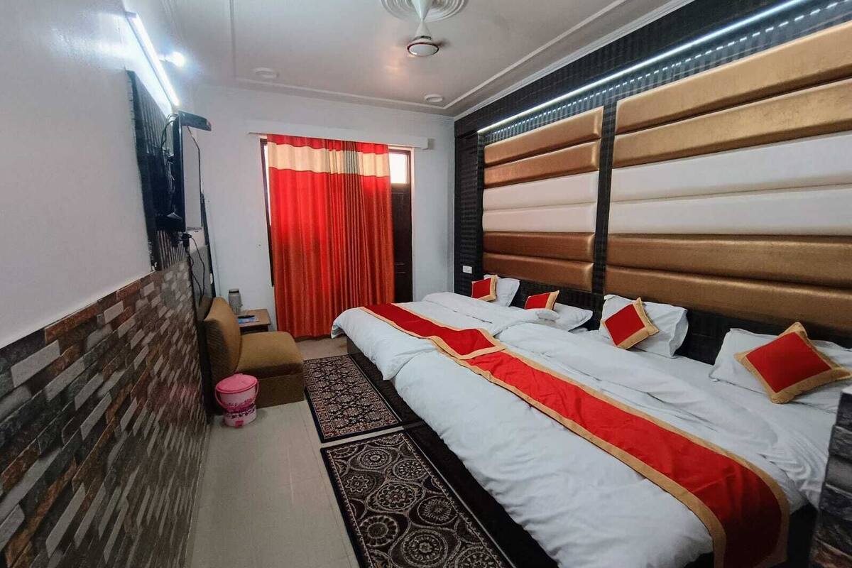 A hotel room with a bed, a chair, a small table, a window with red curtains, and a patterned rug.