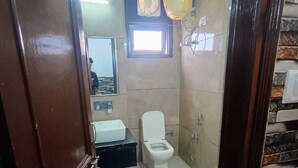 A bathroom with a toilet, sink, and mirror. There is a window above the toilet and a showerhead on the wall. - Hotel New Blue Sapphire Katra (Reasi)