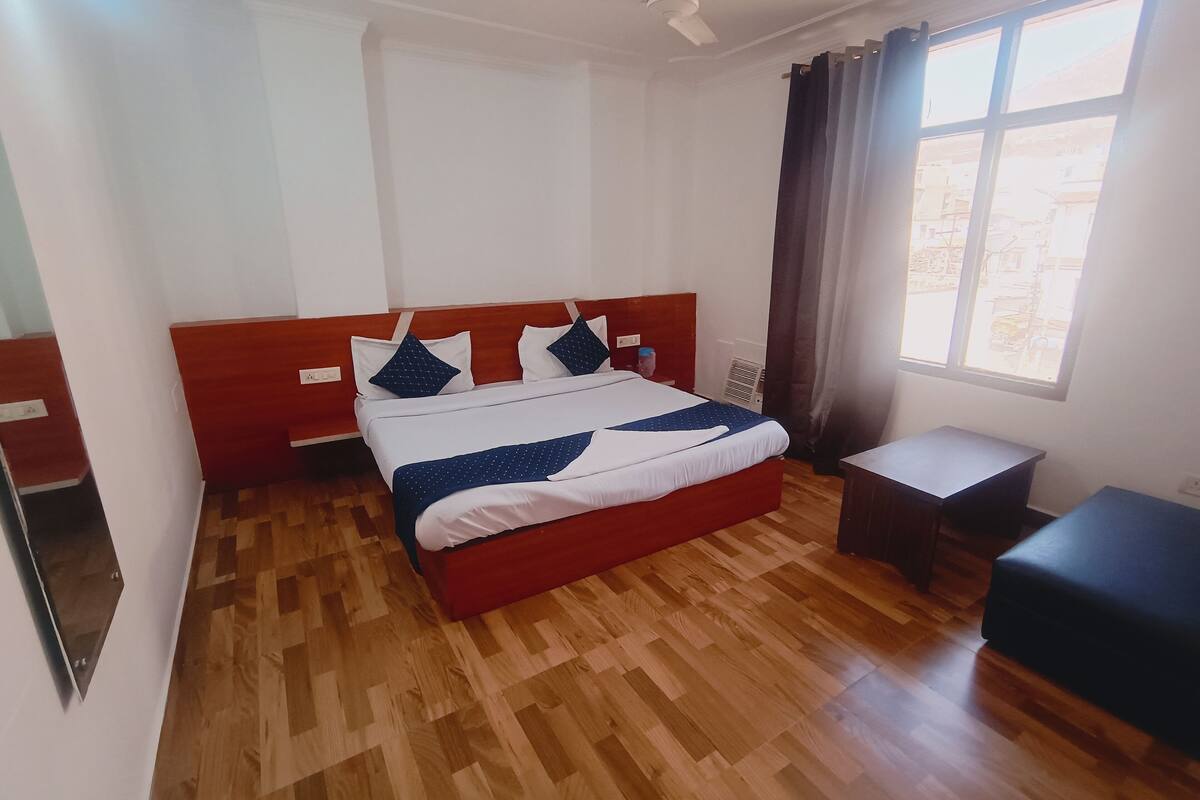 A hotel room with a wooden floor, a bed with white and blue bedding, a small desk, and a window with curtains.