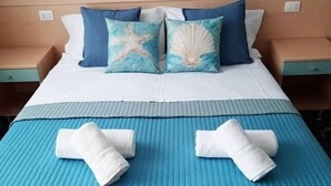 A neatly made bed with a blue bedspread, two pillows with starfish and shell designs, and two rolled towels on the bed. - Residence Rex (Chioggia)