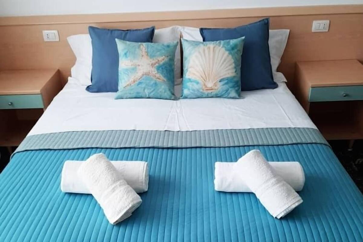 Una cama bien hecha con una colcha azul, dos almohadas con diseños de estrellas de mar y conchas, y dos toallas enrolladas sobre la cama.