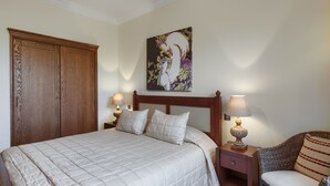 A neatly made bed with a quilted white comforter and two pillows, flanked by wooden nightstands with lamps. A large wooden wardrobe is positioned to the left of the bed. - Aphrodite Hills Rentals - 1 Bedroom Apartment - AHA12 (paphos)