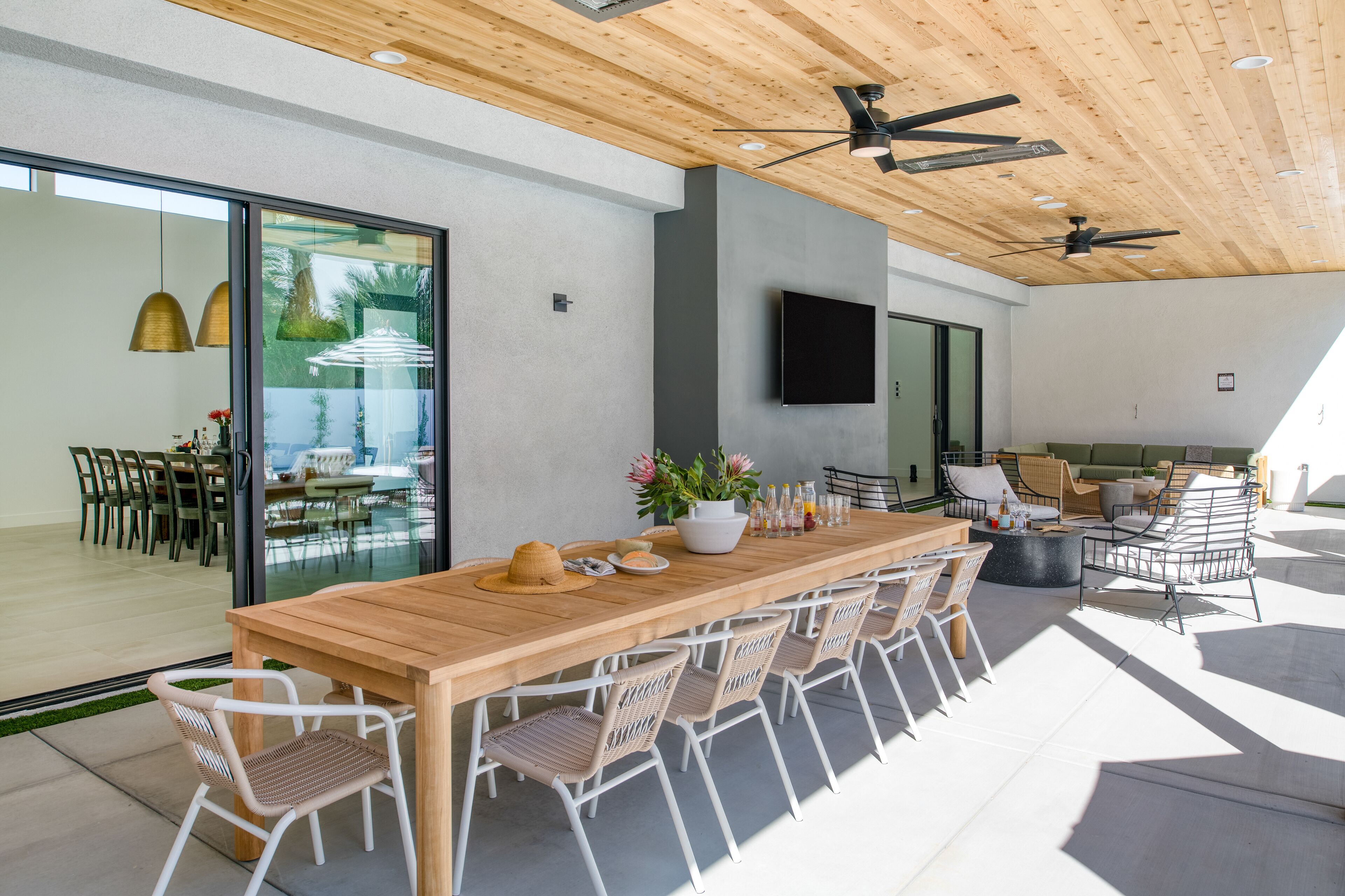 Un vaste espace extérieur aménagé pour les repas, avec une longue table en bois, des chaises en osier blanches et un ventilateur de plafond moderne.