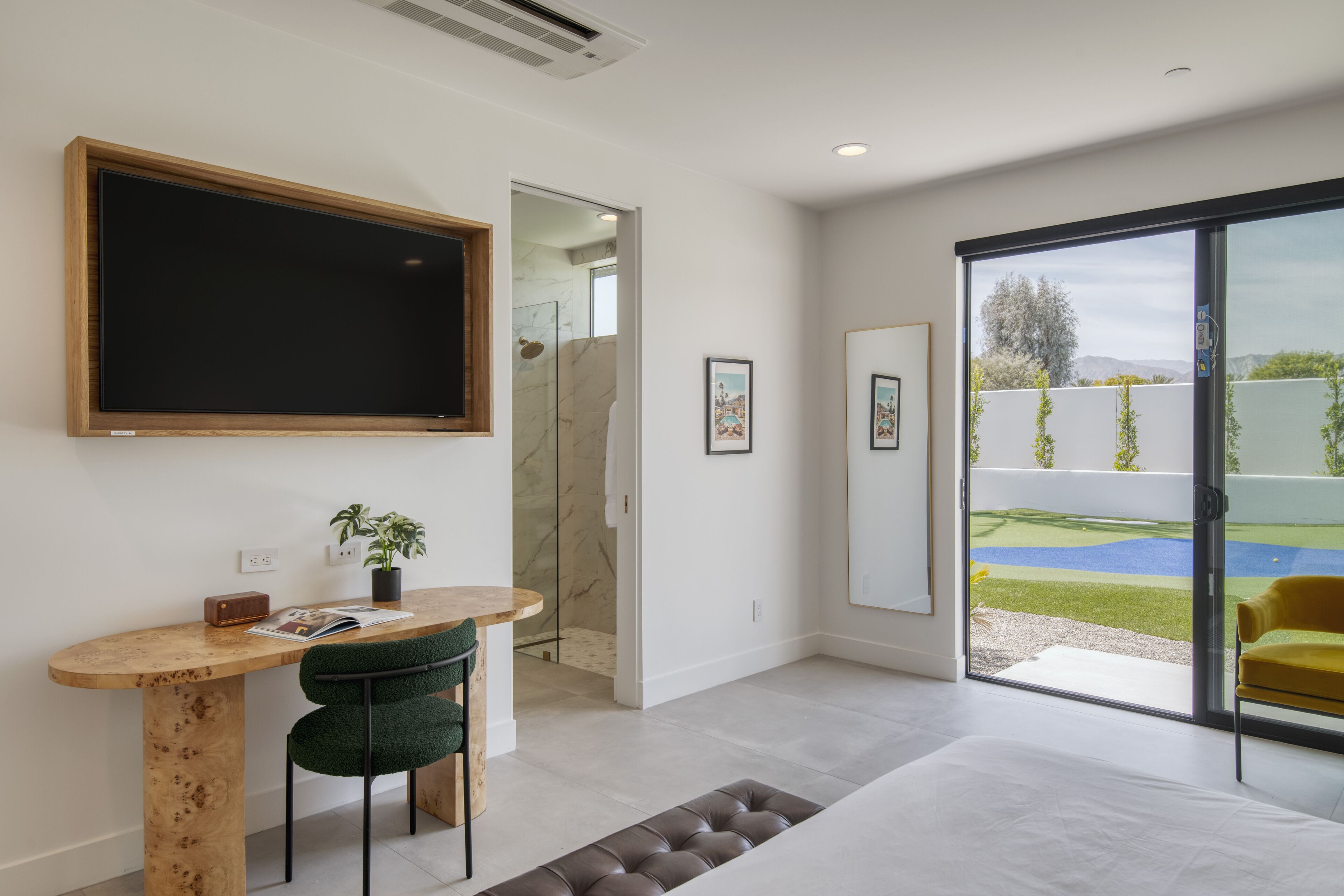 A modern room with a large TV, a wooden desk, a green chair, and a sliding glass door to a garden.