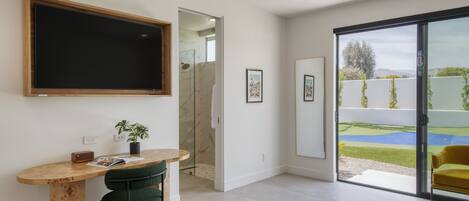 A modern room with a large TV, a wooden desk, a green chair, and a sliding glass door to a garden.