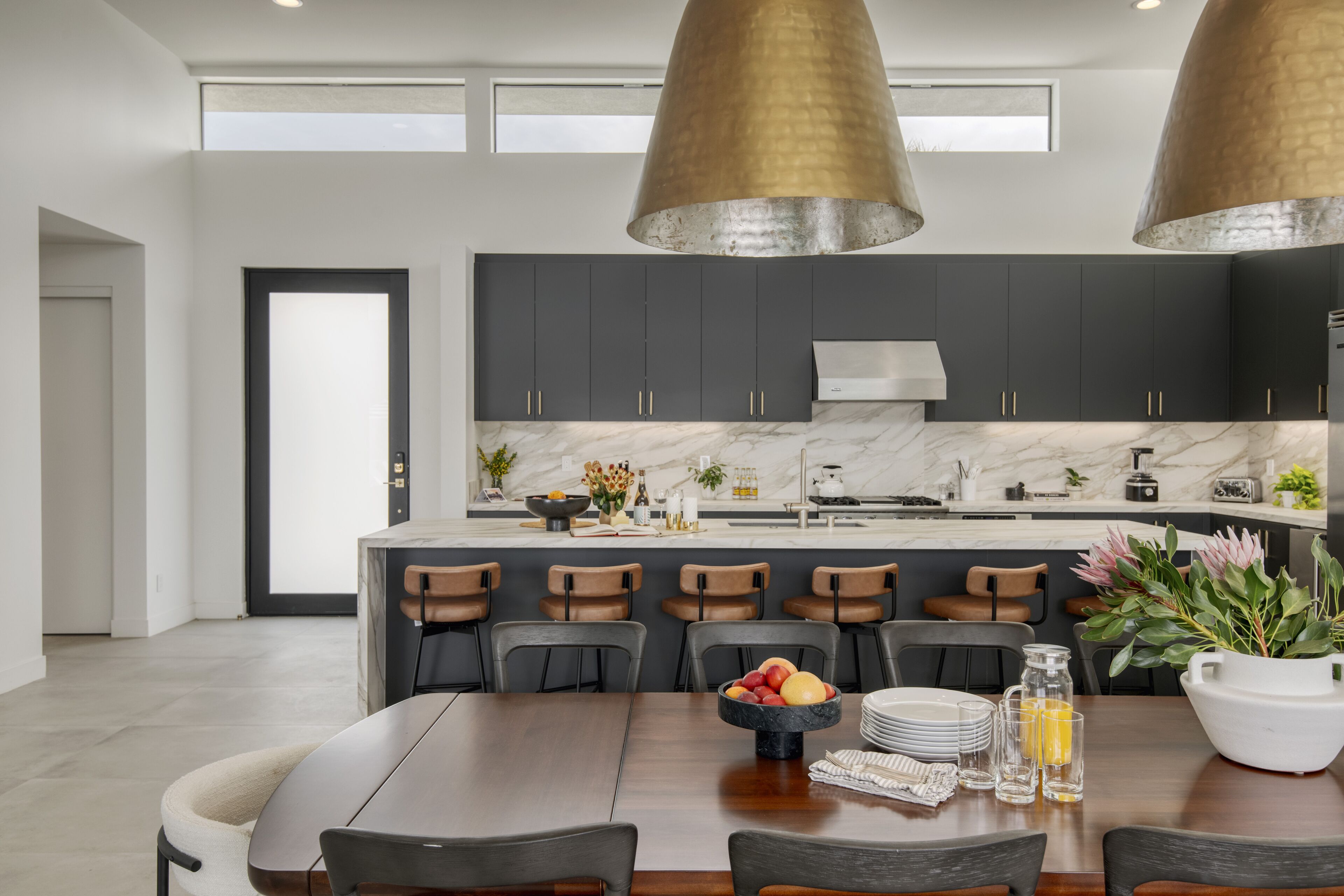 Una cocina moderna con armarios oscuros, encimera de mármol y una isla central. Hay dos lámparas colgantes, una mesa de comedor con sillas y un frutero sobre la isla.