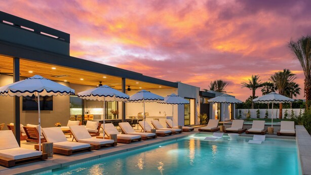 A poolside area with lounge chairs and umbrellas, a modern building with a flat roof, and a sunset sky. - Polo Villas Bella | 2 Homes | Putting Green, Bocce Ball |  Sleeps 32 (La Quinta)