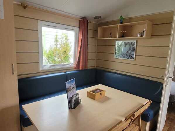 A cozy RV interior with a window, a table with a magazine rack, a cushioned bench, and a shelf with decorative items.