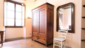 Une pièce avec une armoire en bois, une chaise blanche, un grand miroir et une fenêtre dont les volets sont ouverts.