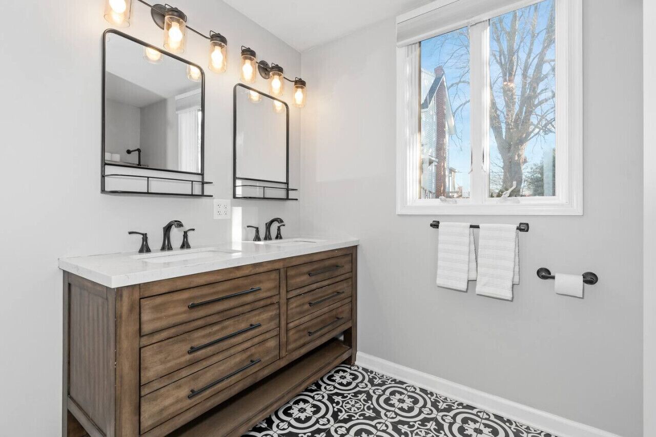 A bathroom with two mirrors, a vanity with drawers, a window with a view of trees, and a patterned floor.