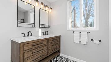 A bathroom with two mirrors, a vanity with drawers, a window with a view of trees, and a patterned floor.