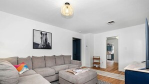 A modern living room with a sectional sofa, a small ottoman, a black and white rug, a framed horse portrait, and a kitchen visible through an open doorway. - White Clay Creek Cottage (Newark)