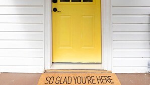 A yellow door with a glass panel, flanked by two black lantern-style lights, and a doormat with the text "SO GLAD YOU'RE HERE" on a porch with white siding. - White Clay Creek Cottage (Newark)