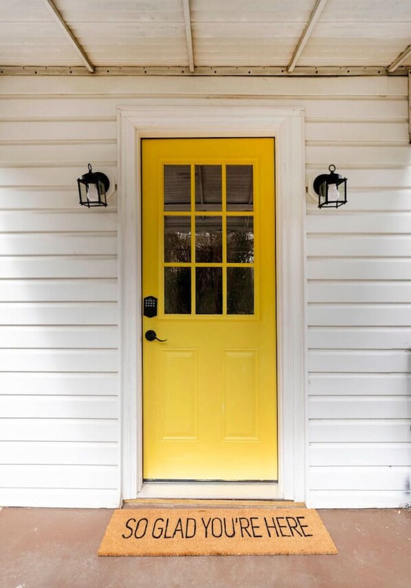 A yellow door with a glass panel, flanked by two black lantern-style lights, and a doormat with the text "SO GLAD YOU'RE HERE" on a porch with white siding.